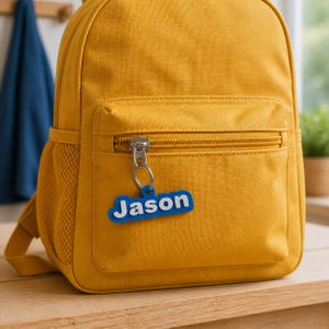 May include: A mustard yellow backpack featuring a front zippered pocket and a side mesh compartment. Attached to the silver metal zipper pull is a blue and white rectangular keychain with the name Jason embossed in white capital letters.