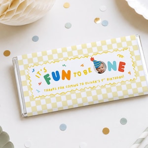 May include: A rectangular chocolate bar wrapper featuring a light yellow and white checkered pattern. The centre label reads IT'S FUN TO BE ONE, THANKS FOR COMING TO OLIVER'S 1ST BIRTHDAY, with a photo of a baby wearing a blue party hat.