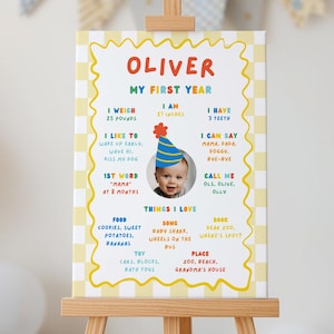 Può includere: Una tavola dei traguardi rettangolare con un bordo a scacchi giallo e bianco che riporta il testo Oliver My First Year. Include sezioni colorate che dettagliano peso, altezza, denti, parole e preferiti, con una foto centrale di un bambino.