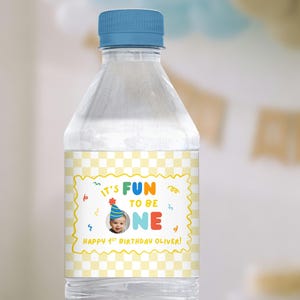 May include: A clear plastic water bottle with a blue cap. The label features a yellow and white checkered pattern and the text "IT'S FUN TO BE ONE" with a photo of a baby. The text "HAPPY 1ST BIRTHDAY OLIVER!" is also on the label.