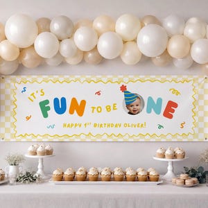 May include: A rectangular birthday banner with a yellow and white checkered border, featuring the text "IT'S FUN TO BE ONE" in colorful letters, a photo of a baby in a party hat, and the text "HAPPY 1ST BIRTHDAY OLIVER!" surrounded by balloons.