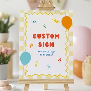 May include: A custom sign displayed on a small wooden easel. The sign features a yellow and white chequered background and a wavy yellow border. The words "CUSTOM SIGN" are in red, with the text "ADD YOUR OWN TEXT HERE!" below. Balloons and confetti are also present.