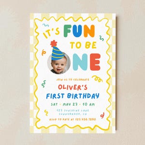 Può includere: Un colorato invito per il primo compleanno con la scritta "It's Fun to be One". L'invito presenta una foto di un bambino con un cappello da festa, circondato da vortici decorativi e il nome "Oliver's First Birthday".
