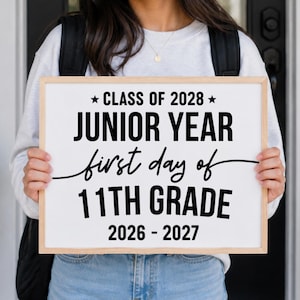 Könnte beinhalten: Ein rechteckiges weißes Schild mit hellem Holzrahmen und schwarzem Text, der CLASS OF 2028, JUNIOR YEAR, first day of 11TH GRADE und 2026 - 2027 lautet. Das Schild wird horizontal gehalten und zeigt eine fette und kursive Typografie.