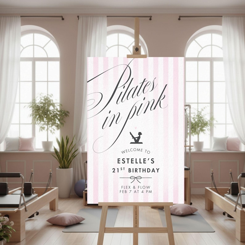 May include: A pink and white striped sign on a wooden easel. The sign reads "Pilates in pink" in elegant script, with additional text below indicating a birthday celebration. The sign is in a bright, airy room with pilates equipment.