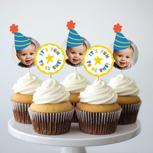 May include: Five cupcakes topped with decorative toppers. Three toppers feature a baby wearing a blue and yellow party hat with a red pom-pom. Two circular toppers feature a yellow star and the text IT'S FUN TO BE ONE! in colorful letters.
