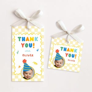 May include: Two gift tags with a yellow checkered background and a white ribbon. The tags say "THANK YOU!" in colorful letters, with a photo of a baby wearing a party hat. The name "OLIVER" is also printed on the tags.