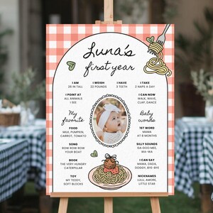 K&ouml;nnte beinhalten: Personalisiertes Schild zum ersten Geburtstag mit einem rosa-wei&szlig; karierten Rand. Das Schild enth&auml;lt ein Foto des Babys und Details &uuml;ber das erste Lebensjahr, wie Gr&ouml;&szlig;e, Gewicht und Lieblingsdinge. Das Schild wird auf einer Holzstaffelei pr&auml;sentiert.