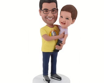 May include: A bobblehead figurine of a man holding a child. The man wears a yellow t-shirt, blue trousers, and glasses. The child wears a light purple top. The base reads "Happy 50th Birthday Padrino".