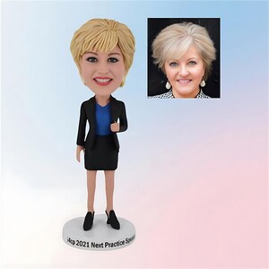 May include: A custom bobblehead figurine of a woman in a business suit, with a matching image of the person. The figurine has a blonde hairstyle, a black blazer, a blue top, and a black skirt. The base of the figurine reads "14cp 2021 Next Practice Spea".