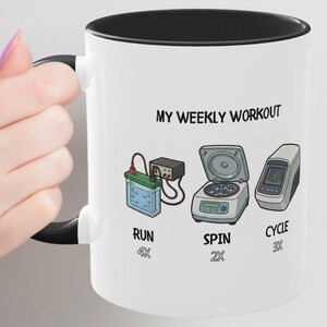Puede incluir: Taza de cerámica blanca con interior y asa negros. La taza presenta el texto "MY WEEKLY WORKOUT" e ilustraciones de equipos de laboratorio con las palabras "RUN", "SPIN" y "CYCLE" debajo. La taza está sostenida por una persona.
