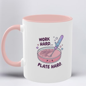Könnte beinhalten: Weißer Keramikbecher mit rosa Innenraum und Henkel. Der Becher zeigt eine Cartoon-Illustration einer Petrischale mit einem Schneebesen und dem Text "WORK HARD... PLATE HARD." Blasen und Herzen sind ebenfalls im Design enthalten.