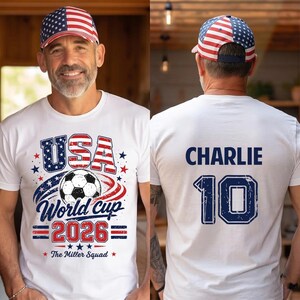 Op de afbeelding: Een wit t-shirt met een afbeelding van een voetbal, de tekst USA World Cup 2026 en The Miller Squad op de voorkant, met de naam CHARLIE en het nummer 10 op de achterkant, gecombineerd met een baseballpet met een Amerikaans vlagpatroon.