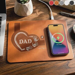May include: A brown leather wireless charging pad with the word "DAD" inside a heart shape, along with the names "Nick", "Lisa", and "Liam". A smartphone is charging on the pad, displaying a 75% charge. A white charging cable is connected.