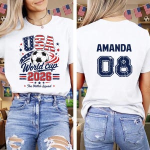 Op de afbeelding: Een wit t-shirt met een afbeelding op de voorkant van een voetbal, de tekst USA World Cup 2026 en The Miller Squad. De achterkant van het shirt toont de naam AMANDA in donkerblauwe letters boven het grote donkerblauwe rugnummer 08.