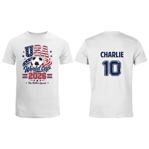 Op de afbeelding: Een wit t-shirt met een afbeelding op de voorzijde van een voetbal, rode en blauwe sterren en de tekst USA World Cup 2026 The Miller Squad. De achterzijde toont de naam CHARLIE in blauwe letters boven het grote blauwe nummer 10.