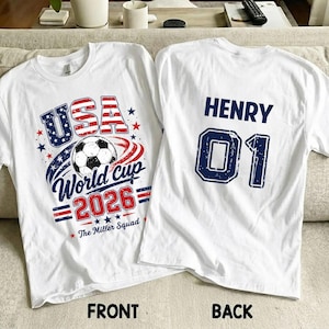 Op de afbeelding: Twee witte t-shirts met een voetbalthema. De voorkant toont een voetbal, rode en blauwe sterren en de tekst USA World Cup 2026 The Miller Squad. De achterkant bevat de naam HENRY en het nummer 01 in grote blauwe blokletters.