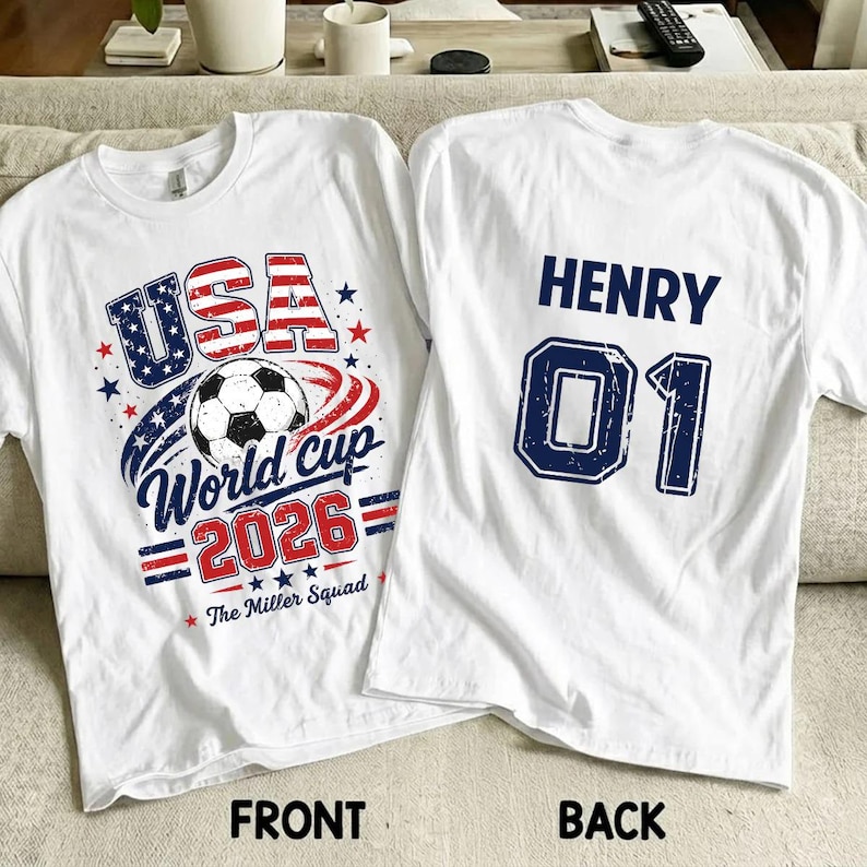 Op de afbeelding: Twee witte t-shirts met een voetbalthema. De voorkant toont een voetbal, rode en blauwe sterren en de tekst USA World Cup 2026 The Miller Squad. De achterkant bevat de naam HENRY en het nummer 01 in grote blauwe blokletters.