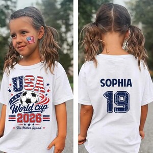 Op de afbeelding: Een wit t-shirt met korte mouwen en een afbeelding op de voorkant met een voetbal, de tekst USA World Cup 2026 en The Miller Squad. De achterkant toont de naam SOPHIA in donkerblauwe letters boven het grote donkerblauwe nummer 19.