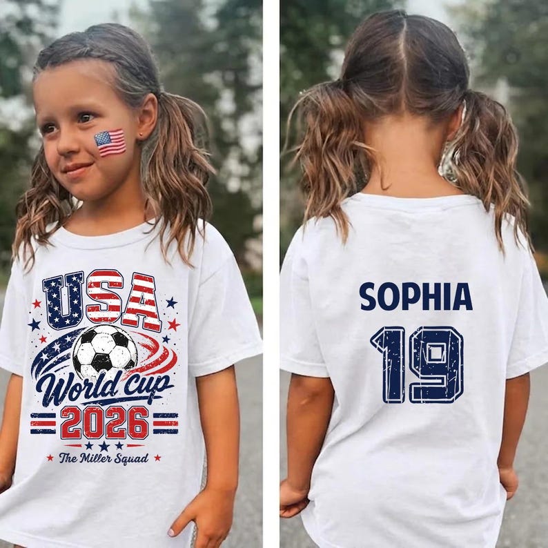 Op de afbeelding: Een wit t-shirt met korte mouwen en een afbeelding op de voorkant met een voetbal, de tekst USA World Cup 2026 en The Miller Squad. De achterkant toont de naam SOPHIA in donkerblauwe letters boven het grote donkerblauwe nummer 19.