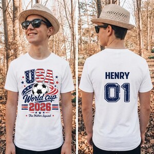 Op de afbeelding: Een wit t-shirt met korte mouwen en een afbeelding op de voorkant met een voetbal, de tekst USA World Cup 2026 en The Miller Squad. De achterkant van het shirt toont de naam HENRY en het nummer 01 in donkerblauwe blokletters.