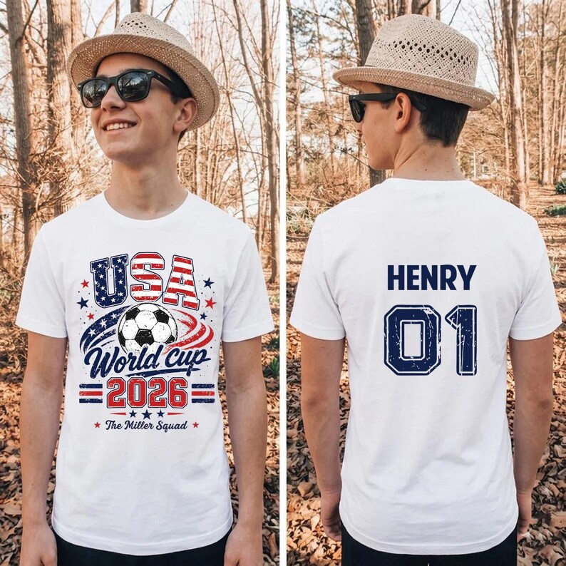 Op de afbeelding: Een wit t-shirt met korte mouwen en een afbeelding op de voorkant met een voetbal, de tekst USA World Cup 2026 en The Miller Squad. De achterkant van het shirt toont de naam HENRY en het nummer 01 in donkerblauwe blokletters.