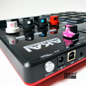 Puede incluir: Primer plano de un controlador MIDI Akai Professional con perillas personalizadas de colores. El controlador es negro con detalles rojos y presenta varios puertos y botones. Las perillas son blancas, negras, azules, rojas y con un diseño rosa y negro.