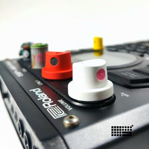 Peut inclure: Gros plan sur un échantillonneur Roland SP-404MKII noir avec des bouchons de peinture en aérosol colorés. Les bouchons sont rouges, blancs et jaunes avec des accents noirs et roses. L'échantillonneur a un bouton de volume et un logo.