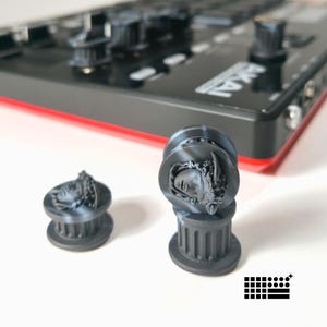 Può includere: Manopole decorative nere e grigie con un design a busto classico. Le manopole sono davanti a un dispositivo elettronico nero con la scritta "AKAI" stampata sopra. Le manopole hanno una base a forma di colonna.