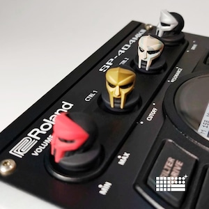 May include: Close-up of a black Roland SP-404MKII sampler with custom knobs featuring various colored mask designs. The knobs are red, gold, silver, and white. The sampler has text that reads "VOLUME", "MIN", "MAX", "CTRL 1", "TRL 2", "CUTOFF", "RESONANCE", "FILTER", and "DRIVE".