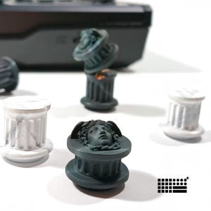 Op de afbeelding: Een verzameling miniatuur, decoratieve kolomvormige objecten. Sommige zijn donkergrijs met een gebeeldhouwd gezicht, andere wit met verticale groeven. De objecten zijn gerangschikt op een wit oppervlak, met een zwart elektronisch apparaat op de achtergrond. Afmetingen in cm.