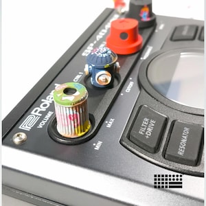 Puede incluir: Primer plano de un sampler Roland SP-404 MKII negro con perillas personalizadas. Las perillas están pintadas con varios diseños, incluyendo una tapa de aerosol roja, una boca de incendios azul y un diseño verde y rosa. El sampler tiene botones etiquetados "FILTER + DRIVE" y "RESONATOR".