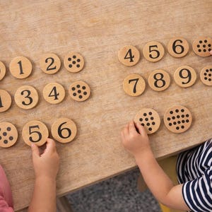 Könnte beinhalten: Holzzahlkacheln mit entsprechenden Punktmustern sind auf einer hellen Holzoberfläche angeordnet. Zwei Kinder interagieren mit den Kacheln, halten und ordnen sie an. Die Zahlen 0-9 sind sichtbar, zusammen mit Punktmustern.