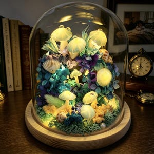 May include: A decorative glass dome with an illuminated miniature mushroom and floral arrangement. The arrangement includes seashells, moss, and colourful flowers, set on a wooden base. The mushrooms glow with a soft light.