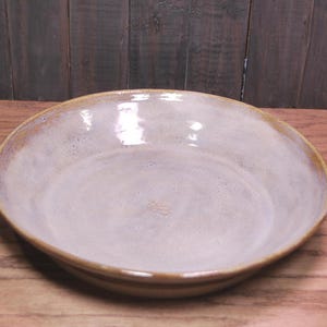 May include: A shallow, round ceramic bowl with a glossy, light lavender-grey interior glaze. The rim and exterior edge feature a contrasting warm, earthy brown tone with a smooth, reflective finish and a slightly uneven, hand-crafted texture.