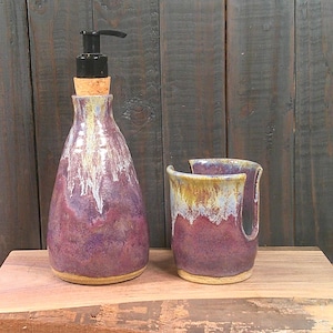 May include: A ceramic soap dispenser with a black pump and cork stopper, paired with a matching open-sided ceramic tumbler. Both pieces feature a mottled purple, white, and yellow glaze with a smooth, rounded shape and a natural tan base.