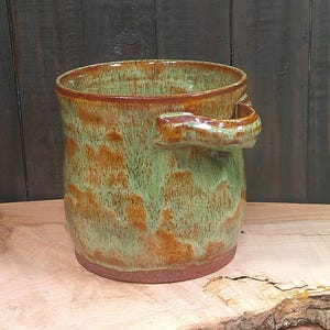May include: A cylindrical ceramic mug featuring a mottled glaze in shades of sage green and burnt orange. The vessel has a thick, sturdy handle attached to the side and a smooth, unglazed reddish-brown base that contrasts with the glossy finish.