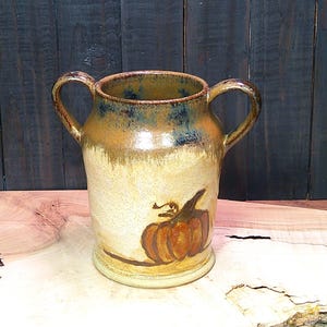 May include: A ceramic vase with two handles, featuring a pumpkin design. The vase has a beige base with brown and blue accents near the rim. The pumpkin is orange with a brown stem and vine.