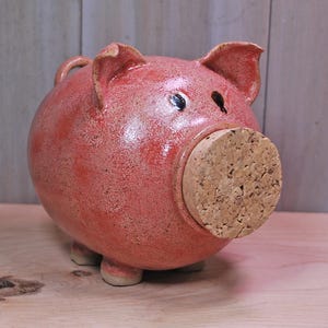 May include: A ceramic piggy bank in a reddish-pink hue, featuring a cork stopper in place of a snout. The bank has small, rounded feet and a loop on top. The surface has a speckled texture, and the eyes are painted black.