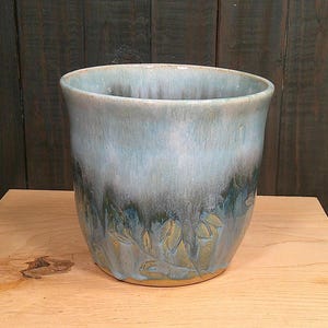 May include: A cylindrical ceramic planter featuring a reactive glaze that transitions from a pale, icy blue at the rim to a deep, forest green at the base. The lower section displays a subtle, embossed botanical leaf pattern in a muted gold tone.