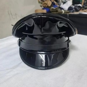 Puede incluir: Un sombrero negro brillante con una corona redondeada, una visera plana y curvada, y una banda alrededor de la base. El sombrero tiene un aspecto brillante, similar al charol. El sombrero está sobre una superficie blanca.
