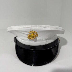 Pode incluir: Um chapéu militar branco com viseira preta e emblema dourado. O chapéu apresenta uma coroa branca e uma aba preta brilhante. Um emblema dourado está centrado na frente do chapéu.