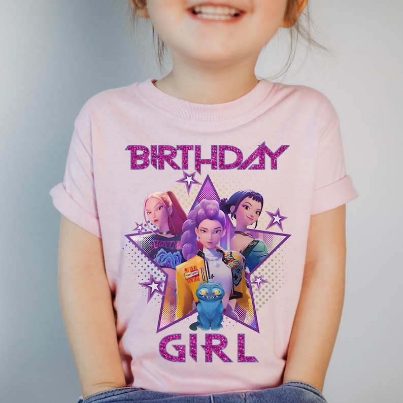 May include: A light pink t-shirt featuring a graphic of three animated girls inside a purple star. The words "BIRTHDAY GIRL" are printed above and below the graphic in glittery pink letters. The shirt has short sleeves.