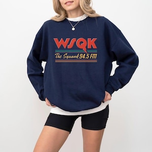 May include: Navy blue sweatshirt with the text "WSQK The Squawk 94.5 FM" in red, orange, and green. The sweatshirt has long sleeves and a crew neck. The person is wearing a white undershirt and black shorts.