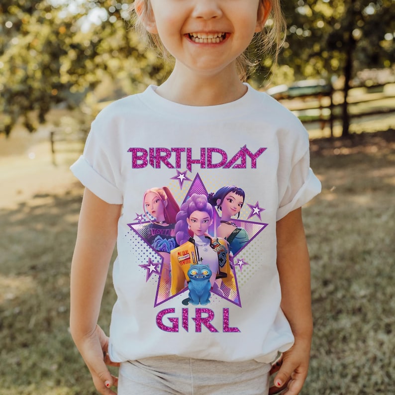 May include: White t-shirt with a graphic featuring three cartoon characters inside a purple star, with the words "BIRTHDAY GIRL" in sparkly pink letters. The shirt is worn by a young child.