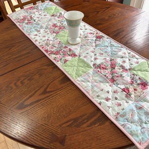 May include: A floral patchwork table runner with pink, green, and blue patterns. The runner features a variety of floral and paisley prints, with a pink border. A white vase with gold trim sits on the runner, adding a decorative touch.
