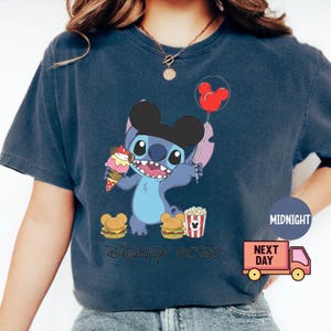 A person wearing a blue t-shirt with a graphic of a character from the Disney movie Lilo and Stitch, holding a balloon and a Mickey Mouse balloon.