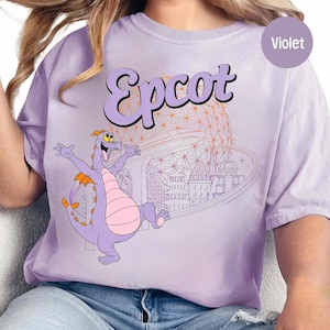 May include: Violet-colored t-shirt featuring the word "Epcot" in a stylized font, with a cartoon dragon character and a graphic of a theme park. The shirt has a relaxed fit.
