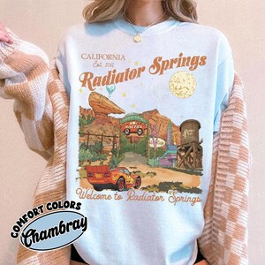 a person wearing a white t-shirt with a graphic design depicting a desert scene with a car, a water tower, and a sign that says "Radiator Springs".