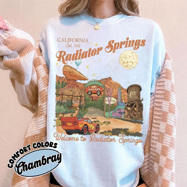 a person wearing a white t-shirt with a graphic design depicting a desert scene with a car, a water tower, and a sign that says "Radiator Springs".